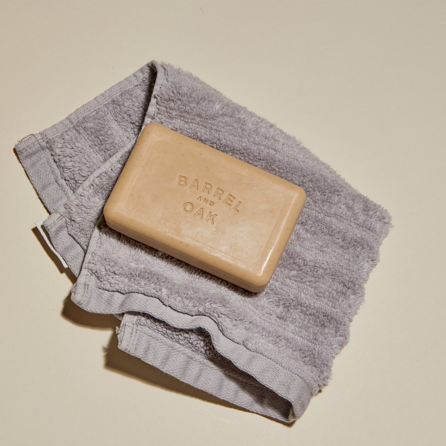 A beige Exfoliating Soap Bar - Spiced Sandalwood 6 oz sits on a folded gray washcloth against a light background, enriched with kaolin clay for gentle exfoliation.
