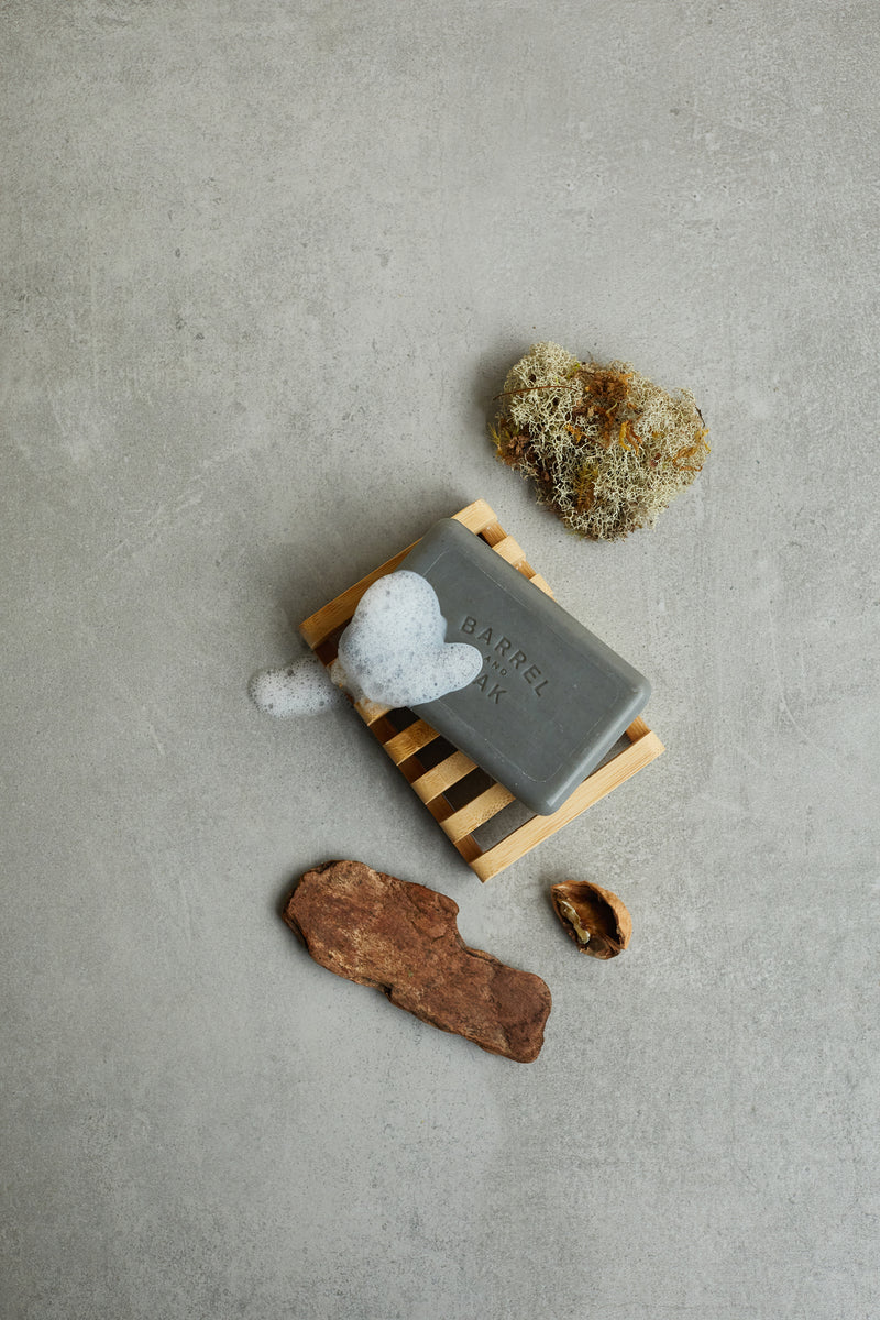 The Exfoliating Soap Bar - Black Oak 6 oz. rests on a wooden dish with foamy lather, surrounded by moss and two brown stones, all arranged on a gray surface.