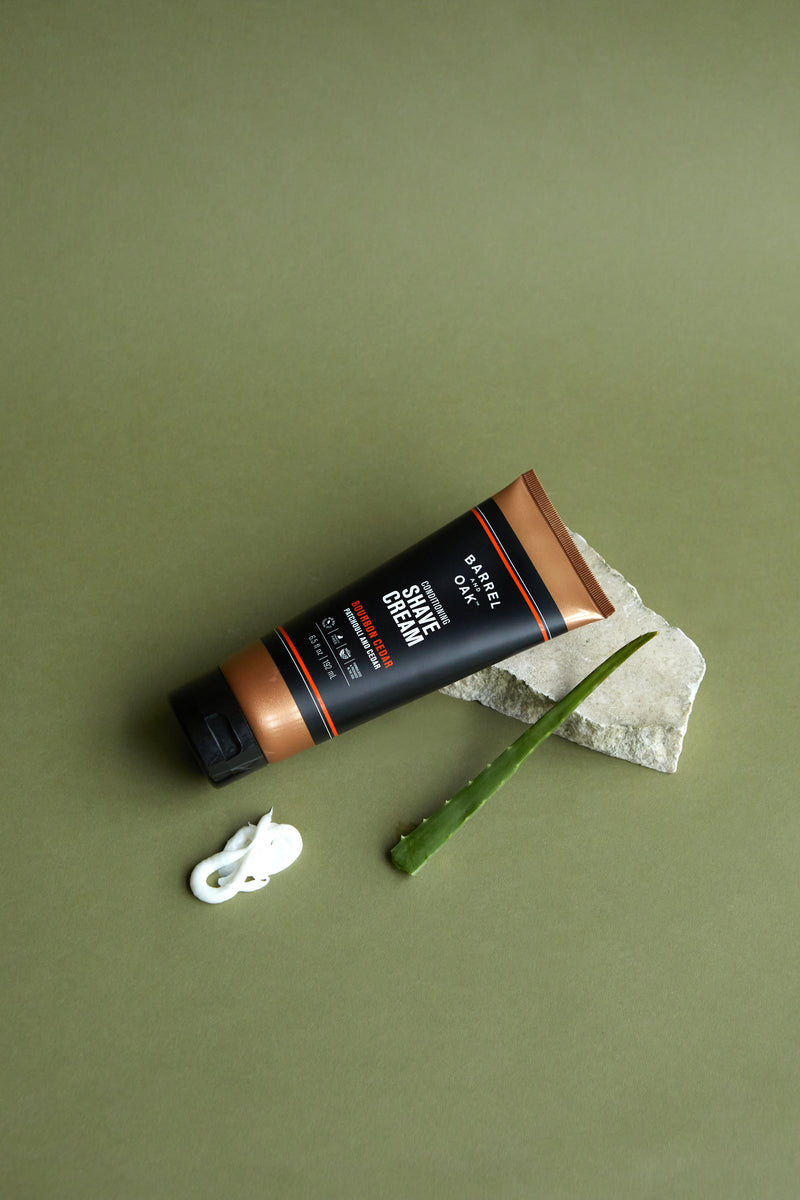 A tube of Conditioning Shave Cream - Bourbon Cedar 6.5 fl oz. rests on stone beside an aloe vera leaf and a dollop of cream, all set against a green background—infused with nourishing hemp seed oil for extra hydration.