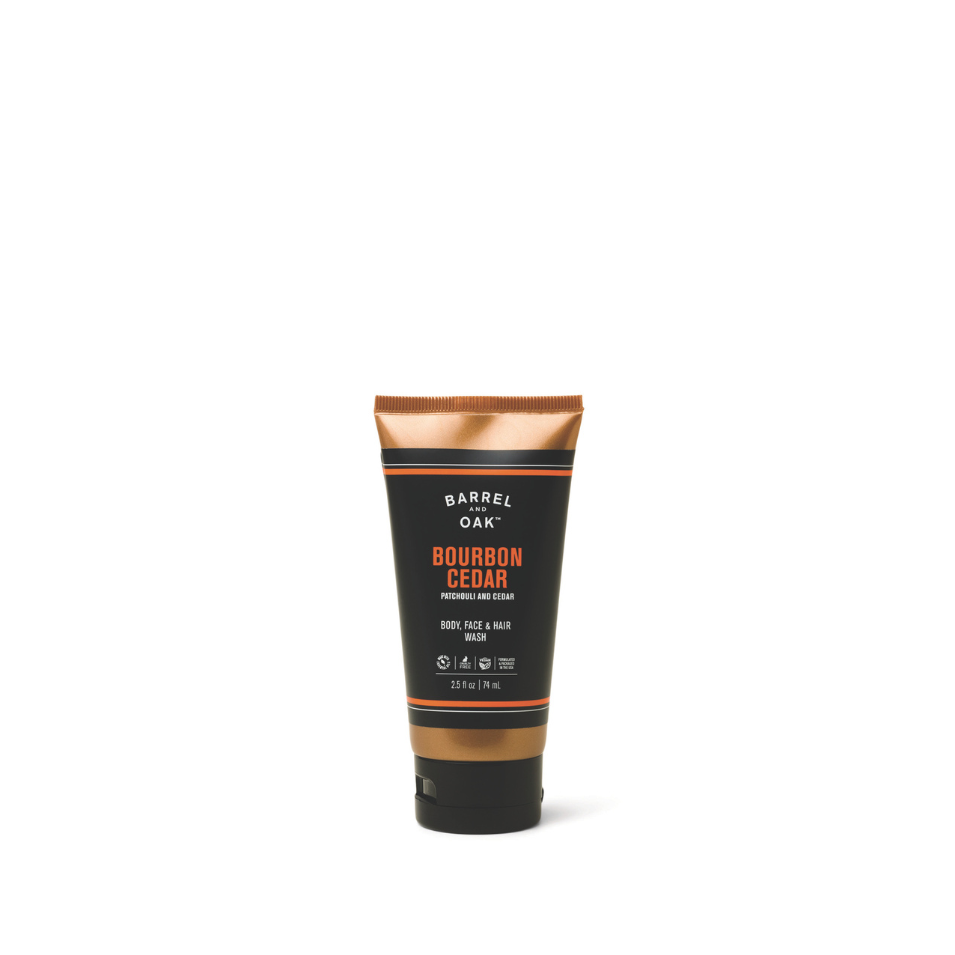 A 2.5 fl oz tube of Barrel & Oak Hair, Face, and Body All In One Wash - Bourbon Cedar is centered on a white background. The black tube with copper accents displays white and orange text for an all-in-one men's wash.
