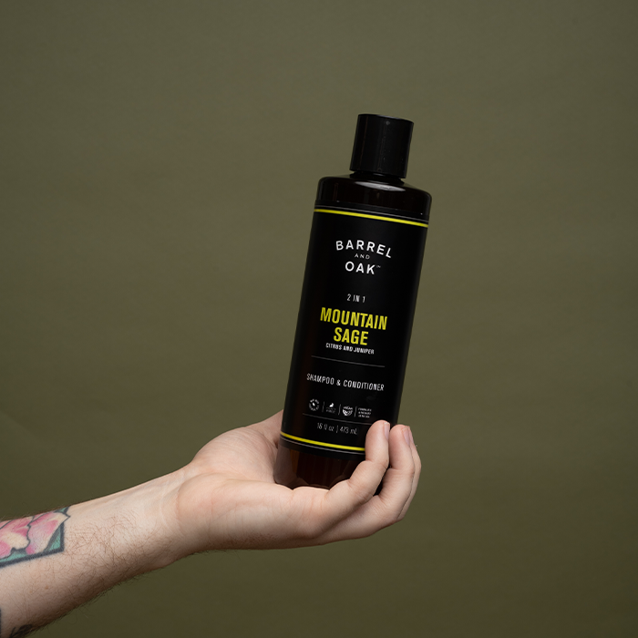 A tattooed hand holds a black bottle of Barrel Oak 2-in-1 Mountain Sage Extra Strength Shampoo & Conditioner against an olive green background.