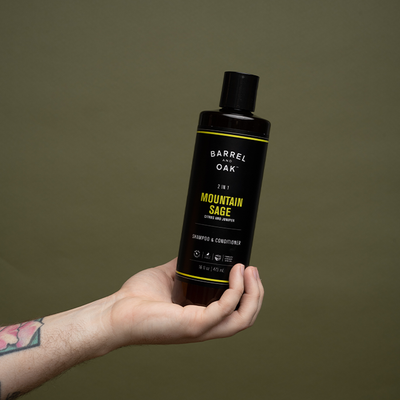 A tattooed hand holds a black bottle of Barrel Oak 2-in-1 Mountain Sage Extra Strength Shampoo & Conditioner against an olive green background.