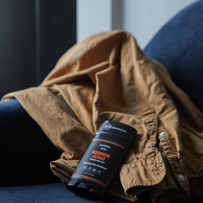 A stick of Aluminum Free Deodorant - Bourbon Cedar 2.7 oz. sits on a brown button-up shirt, which is draped over a dark blue chair.