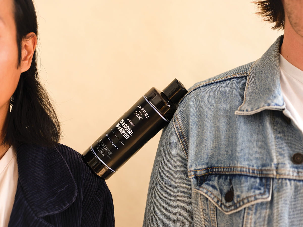 A bottle of Cleansing Charcoal Shampoo - Mint & Eucalyptus 16 oz. perches between two people—one in a dark jacket, the other in denim—with only their heads and upper bodies partially visible.