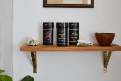 A wooden shelf displays three black jars, including Aluminum Free Deodorant - Spiced Sandalwood 2.7 oz., alongside two small towels and a brown bowl.