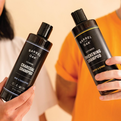 Two people hold up bottles of Barrel and Oak shampoo; one is Cleansing Charcoal Shampoo and the other is Caffeinated Thickening Shampoo. Only their torsos are visible, one in white and one in orange clothing.
