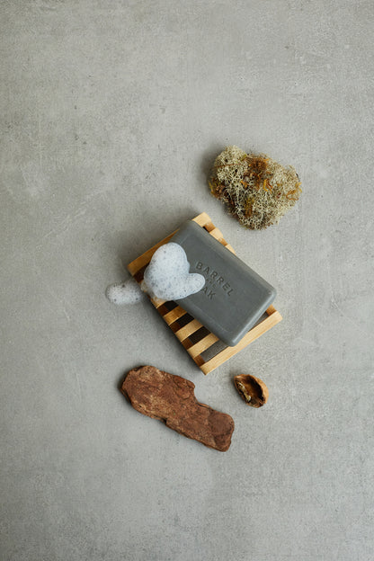 The Exfoliating Soap Bar - Black Oak 6 oz. rests on a wooden dish with foamy lather, surrounded by moss and two brown stones, all arranged on a gray surface.