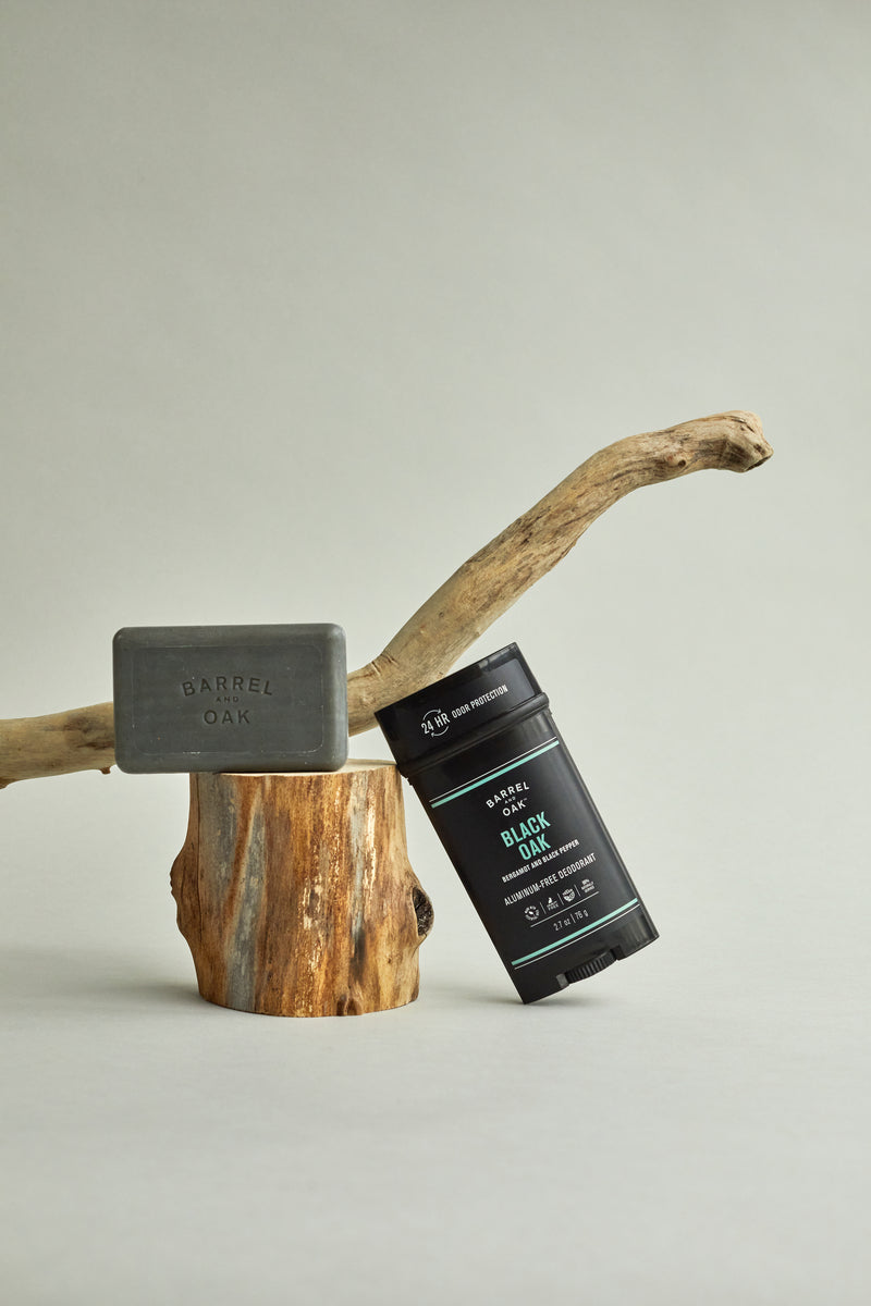 A black soap bar labeled Barrel & Oak sits on a tree stump next to a black Aluminum Free Deodorant - Black Oak 2.7 oz., set against a neutral background with driftwood, highlighting the vegan deodorant’s Bergamot and Black Pepper scent.
