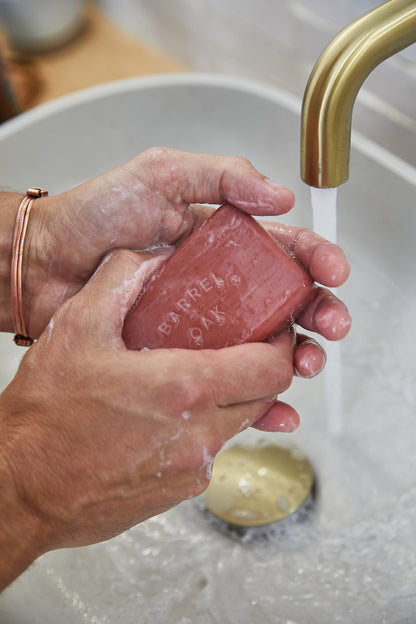 A person lathers the Exfoliating Soap Bar - Bourbon Cedar 6 oz. under running water from a gold faucet above a white sink.
