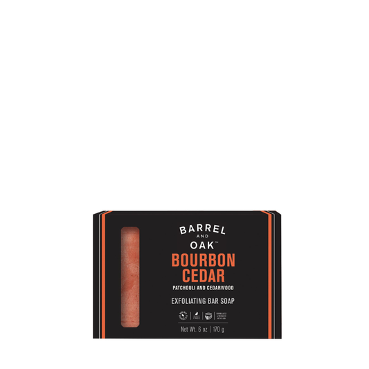 A black box of Exfoliating Soap Bar - Bourbon Cedar 6 oz has a vertical window showing the reddish soap inside. The label notes patchouli, cedarwood, sustainable palm oil, and the weight is 6 oz (170 g).