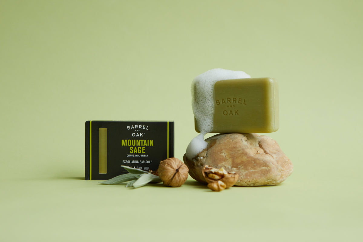 A 6 oz. Exfoliating Soap Bar - Mountain Sage sits foaming on a rock next to its packaging, walnuts, and sage leaves, all against a light green background.