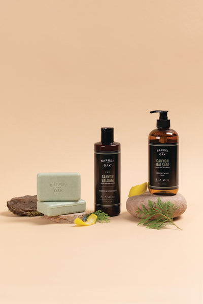 Three Barrel Oak Canyon Balsam personal care products, including a bar soap, a bottle, and a pump bottle, are displayed with a rock, bark, greenery, and lemon peel on a beige background.