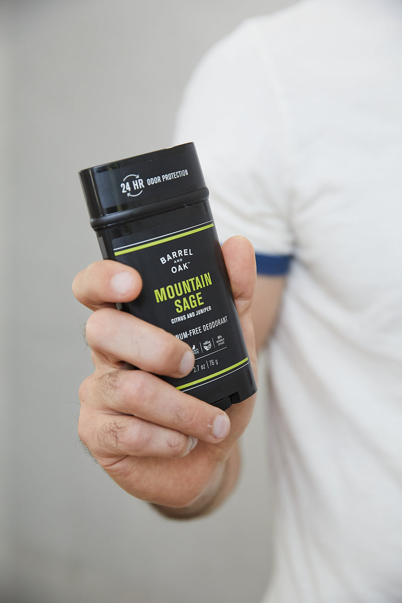 A person in a white t-shirt holds up a black container of Aluminum Free Deodorant - Mountain Sage 2.7 oz., labeled with “24 HR odor protection,” green and yellow text, and highlighting its cruelty-free formula with essential oils.