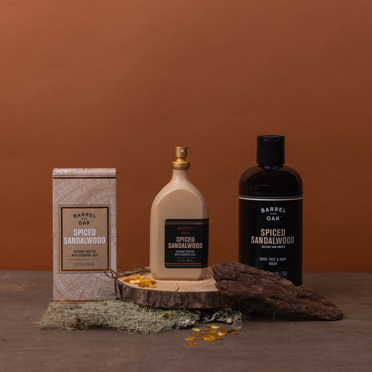 A box, a beige pump bottle, and a black Hair, Face, and Body All In One Wash - Spiced Sandalwood 16 fl oz rest on wood with moss, amber stones, and wood accents against a warm brown background.
