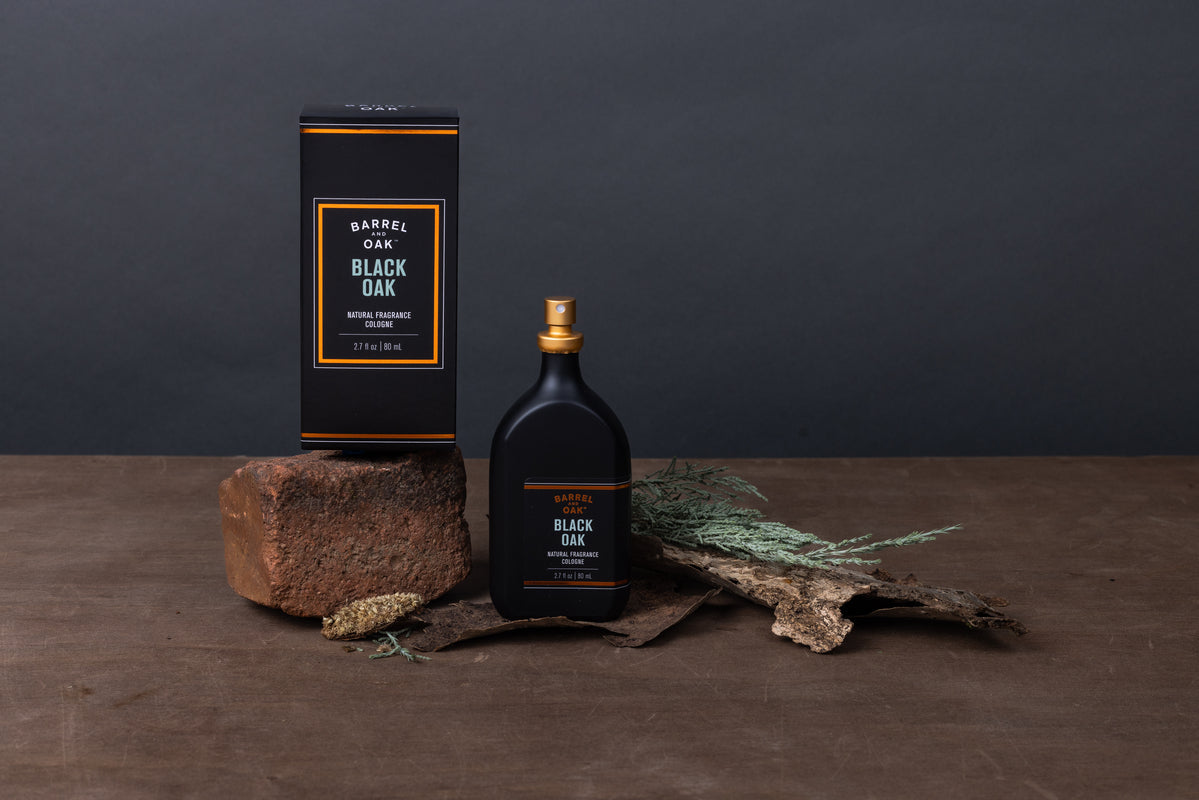 A bottle and box of Natural Fragrance Cologne - Black Oak 2.7 fl oz. are showcased on a brown surface with bark, a brick, green leaves, and a pine cone, emphasizing this earthy essential oil-based scent against a dark gray background.