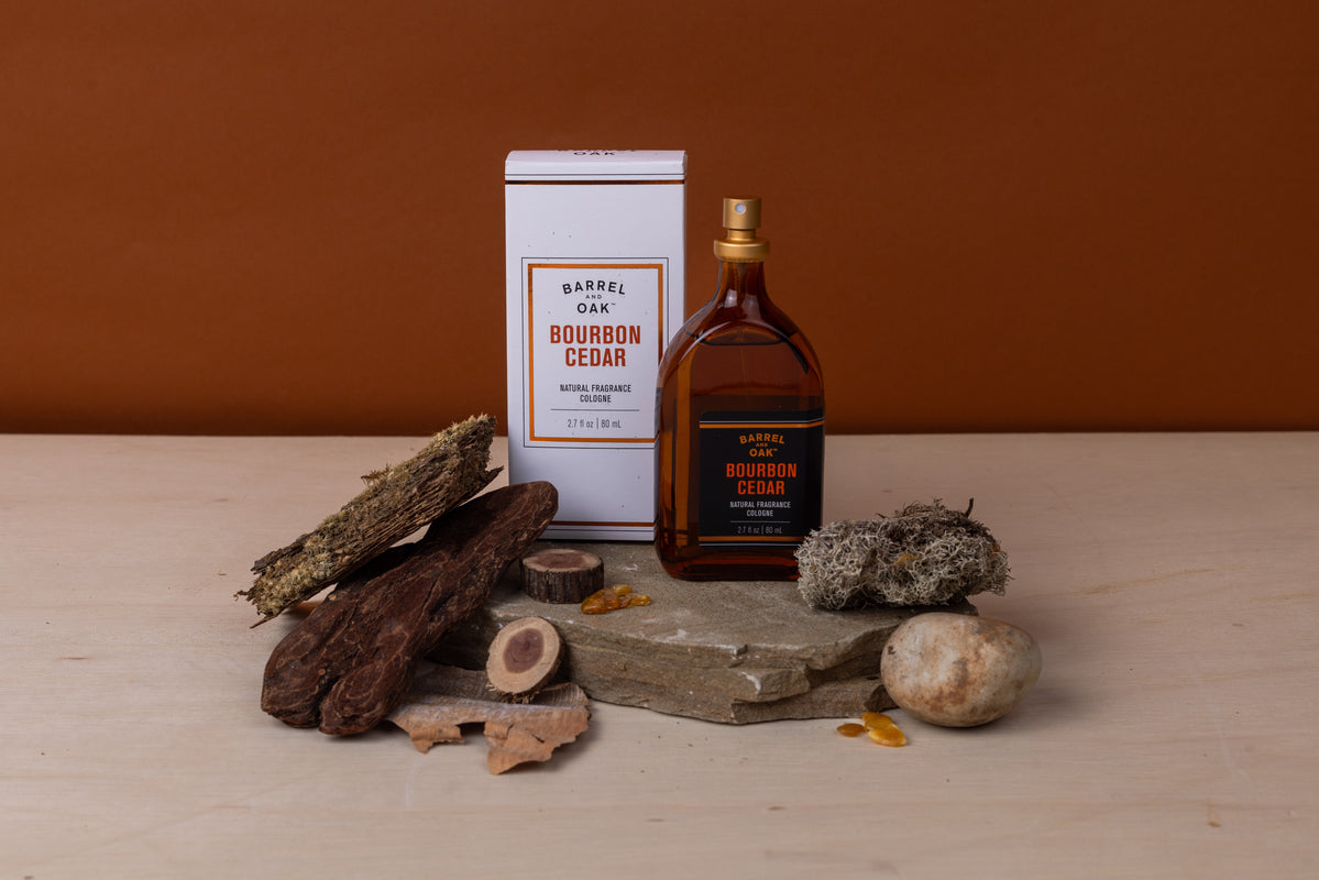 A bottle and box of Natural Fragrance Cologne - Bourbon Cedar 2.7 fl oz. are shown on stone with wood, bark, moss, and amber stones against a brown background, emphasizing the natural essence of this men's cologne.