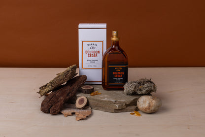 A bottle and box of Natural Fragrance Cologne - Bourbon Cedar 2.7 fl oz. are shown on stone with wood, bark, moss, and amber stones against a brown background, emphasizing the natural essence of this men's cologne.