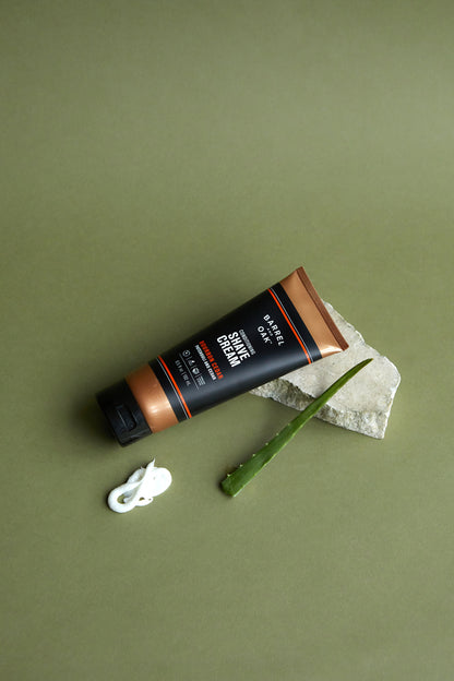 A tube of Conditioning Shave Cream - Bourbon Cedar 6.5 fl oz. rests on stone beside an aloe vera leaf and a dollop of cream, all set against a green background—infused with nourishing hemp seed oil for extra hydration.