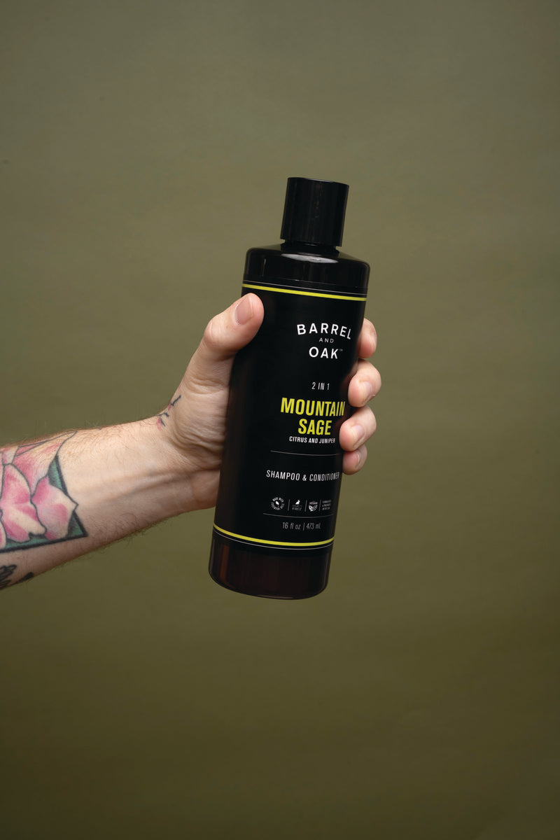 A tattooed hand holds a black bottle of 2 in 1 Shampoo - Mountain Sage 16 fl oz. against a plain olive-green background.