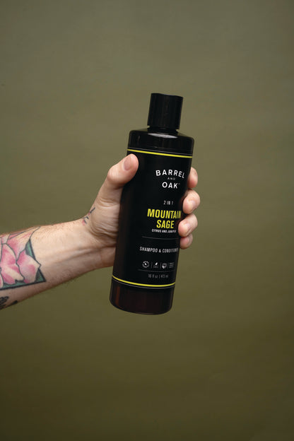 A tattooed hand holds a black bottle of 2 in 1 Shampoo - Mountain Sage 16 fl oz. against a plain olive-green background.