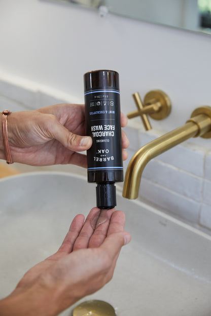 A person squeezes Cleansing Charcoal Face Wash - Mint & Eucalyptus 8 oz. from a black bottle onto their palm over a bathroom sink with a gold faucet.