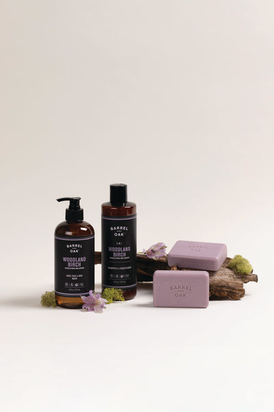 Two bottles of Barrel Oak Woodland Birch body care products stand on a surface beside two lavender soap bars, moss, and purple flowers, with a piece of wood adding a natural touch to the arrangement.
