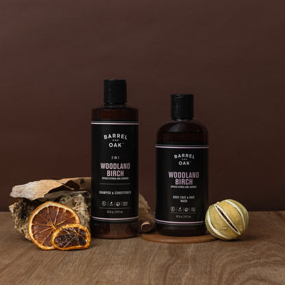 Two bottles of Woodland Birch Hair, Face, and Body All In One Wash rest on a wooden surface with dried orange, birch bark, and a citrus accent, all against a brown background.