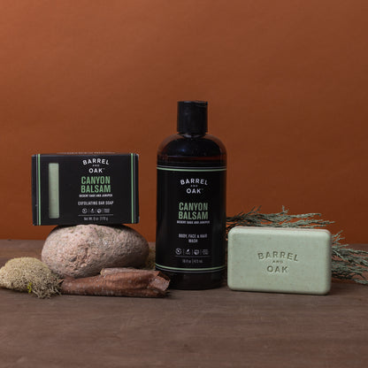 Barrel and Oak's Hair, Face and Body All In One Wash - Canyon Balsam is displayed with natural elements such as rock, stick, moss, and greenery on a brown surface set against a rust-colored background.
