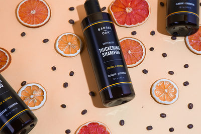 A bottle of Barrel Oak Thickening Shampoo is surrounded by coffee beans and dried citrus slices on a peach-colored background, with additional shampoo bottles partially visible at the edges.