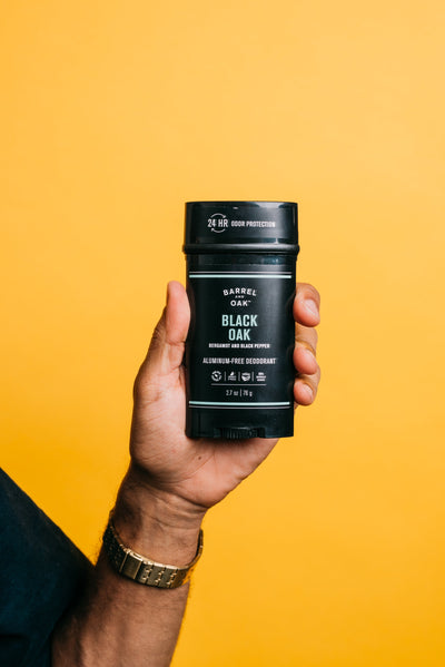 A hand holds a stick of Barrel & Oak Black Oak aluminum-free deodorant against a solid yellow background. The label highlights 24-hour odor protection.